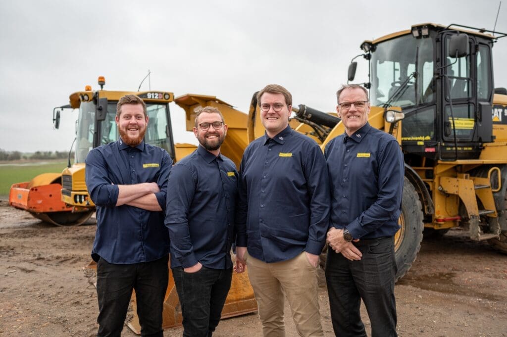 Fire smilende mænd fra IvanBJ i blå skjorter foran gule CAT entreprenørmaskiner og en vejtromle på en byggeplads.