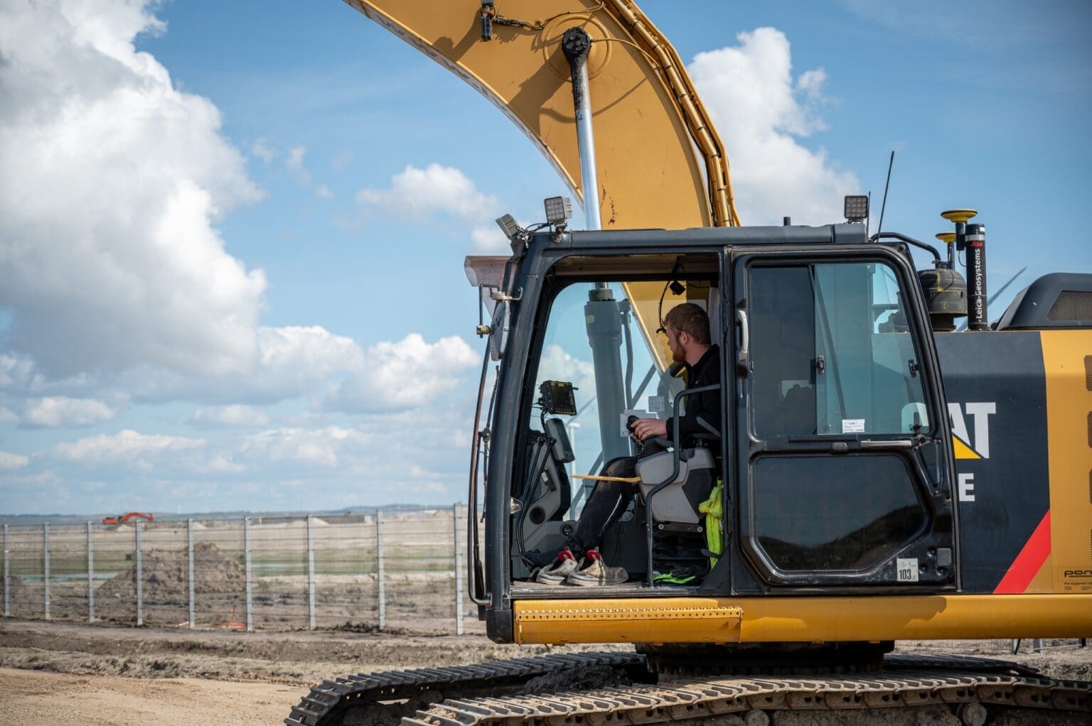 Maskinfører i gul og sort CAT gravemaskine på byggeplads. Manden betjener maskinen i kabinen under åben himmel.