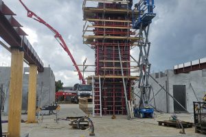 Arbejdere på stillads og lift ved høj betonsøjle under opførelse med rød forskalling og betonpumpe på byggeplads.