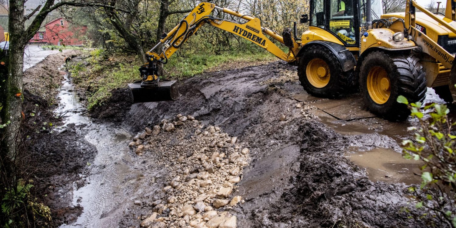 Gul Hydrema gravemaskine rydder op i et mudret vandløb med sten og jord. Maskinen står på en mudret byggeplads.