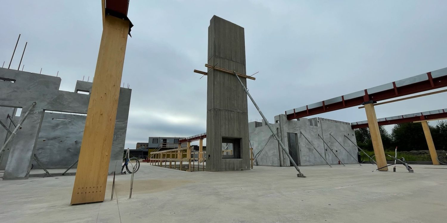 Oversigtsbillede af byggeplads med delvist opførte betonvægge, store træsøjler der bærer stålbjælker under grå himmel.