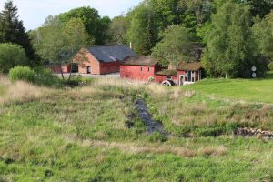 Oversigtsbillede af en rød vandmølle med vandhjul, røde og murstensbygninger, et vandløb og grønne marker omgivet af træer.