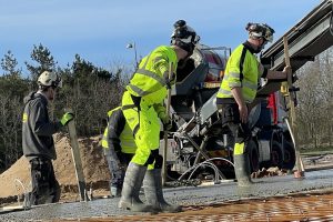 Tre byggearbejdere i sikkerhedsudstyr udfører betonarbejde med en betonbil i baggrunden. Armeringsjern i forgrunden.
