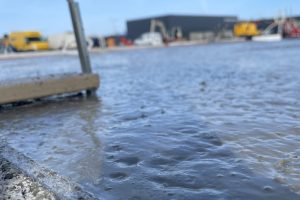 Nærbillede af friskstøbt våd beton med grus langs kanten på en byggeplads under blå himmel. Slørede maskiner i baggrunden.