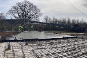 Byggearbejdere støber et fundament med betonpumpe og lastbil. Armering og rør er synlige under den våde beton.