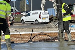 To arbejdere i sikkerhedsveste støber beton på en byggeplads med en betonpumpe og en hvid varevogn fra IvanBJ i baggrunden.