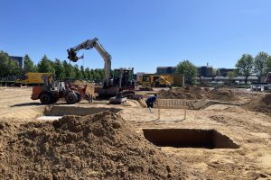 En byggeplads med en gravemaskine, en Atlas gummiged, jordbunker og en arbejder i gang med gravearbejde.