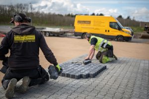 To entreprenører fra Ivan Bastrup Jakobsen lægger mørkegrå belægningssten på en byggeplads. Gul varevogn i baggrunden.